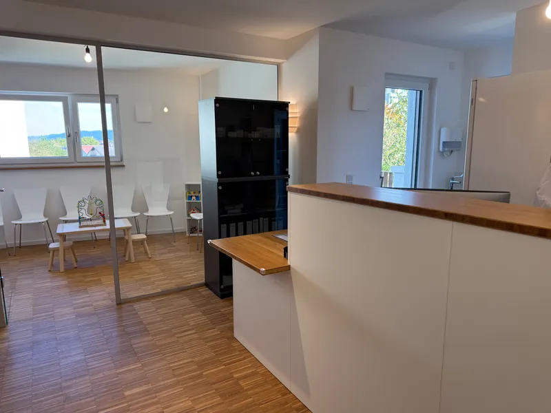 Empfangstresen mit Holzplatte, dahinter das verglaste Wartezimmer